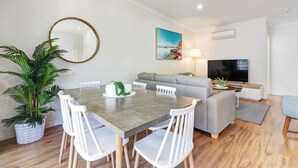 A modern living room with a grey sofa, a flat-screen TV, a dining table with white chairs, and a large potted plant.