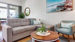 A modern living room with a sofa, armchair, round coffee table, and a painting of a coastal scene.