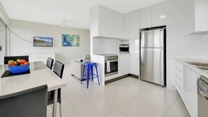 A modern kitchen with white cabinetry, stainless steel appliances, and a dining area with a white table and blue stool.