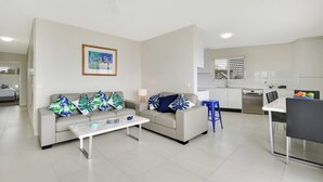 A modern living room with a sofa and coffee table, a dining area with a table and chairs, and a kitchen with white cabinets and appliances.