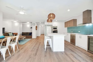 A modern kitchen with white cabinetry, a central island, and a dining area with white chairs. - Lahaina 4 Beachfront Apt Mooloolaba (Maroochydore)