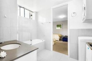 A modern bathroom with a bathtub, a sink, and a toilet. There is a doorway leading to a bedroom with a bed and a blue chair. - Headlands 16 Ocean View Holiday (Alexandra Headland)