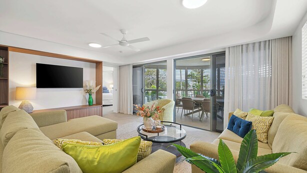 A modern living room with a flat-screen TV, comfortable seating, a ceiling fan, and a glass door leading to an outdoor area. - Riverdance 05 Riverfront Apt (Mooloolaba)