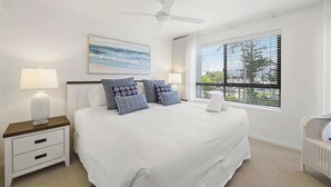 A bedroom with a bed, nightstand, lamp, ceiling fan, window with blinds, and a painting on the wall. - Riverdance 05 Riverfront Apt (Mooloolaba)