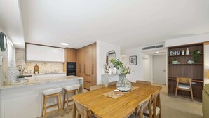 A modern kitchen with white cabinets, a marble countertop, and wooden chairs around a central dining table. - Riverdance 05 Riverfront Apt (Mooloolaba)