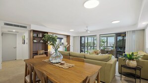 A modern living room with a wooden dining table, beige sofas, a bookshelf, and a large window with a view. - Riverdance 05 Riverfront Apt (Mooloolaba)