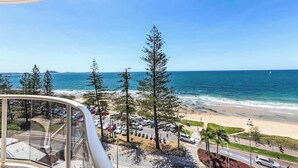 Blick aufs Meer von einem Balkon mit Strand, geparkten Autos und hohen Bäumen.
