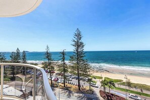 Vista costera desde un balcón con playa, coches aparcados y altos árboles.