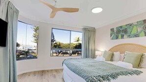 Quarto com cama, janelas amplas, ventilador de teto e TV fixada na parede.