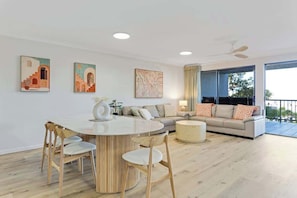 A modern living room with a round dining table, wooden chairs, a sofa, a coffee table, and artwork on the walls. - Riverdance 06 Riverfront Apt (Mooloolaba)