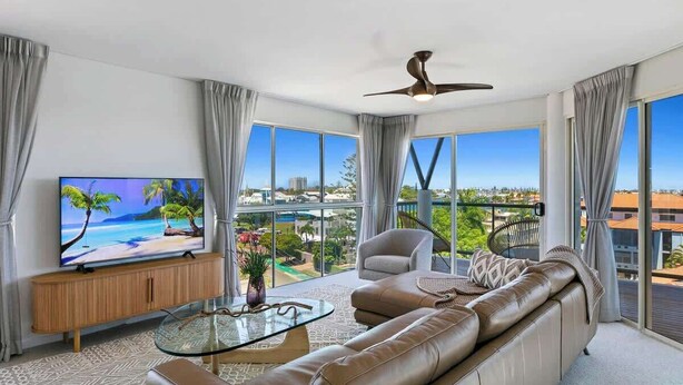 A modern living room with a large TV, leather sofa, and a balcony with a city view. - Luxurious Penthouse Near Beach with Unmatched Views (Mooloolaba)