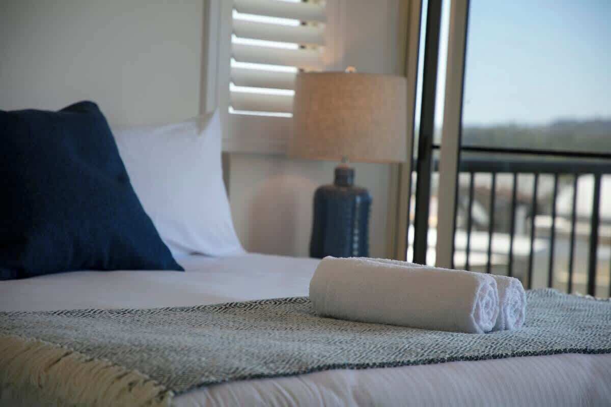 A bed with a rolled towel, a dark pillow, a lamp, and a view through a window.