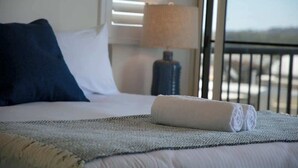 A bed with a rolled towel, a dark pillow, a lamp, and a view through a window. - Luxurious Penthouse Near Beach with Unmatched Views (Mooloolaba)