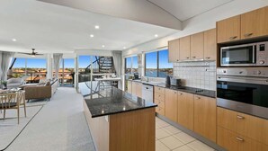 A modern kitchen with wooden cabinets, stainless steel appliances, a central island, and a view of the city. - Luxurious Penthouse Near Beach with Unmatched Views (Mooloolaba)