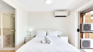 A hotel room with a bed, a nightstand, a lamp, an air conditioning unit, and a sliding glass door leading to a balcony.