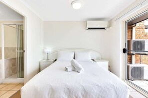 Habitación de hotel con cama, mesita de noche, lámpara, equipo de aire acondicionado y puerta corrediza de vidrio que da a un balcón.