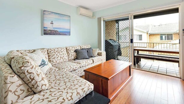 A living room with a sofa, a coffee table, a painting of a lighthouse, and a balcony with a view of houses.