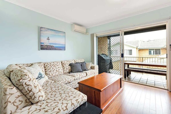 A living room with a sofa, a coffee table, a painting of a lighthouse, and a balcony with a view of houses.