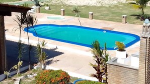A rectangular swimming pool with a clear blue water feature, surrounded by a brick patio, palm trees, and a view of the beach.