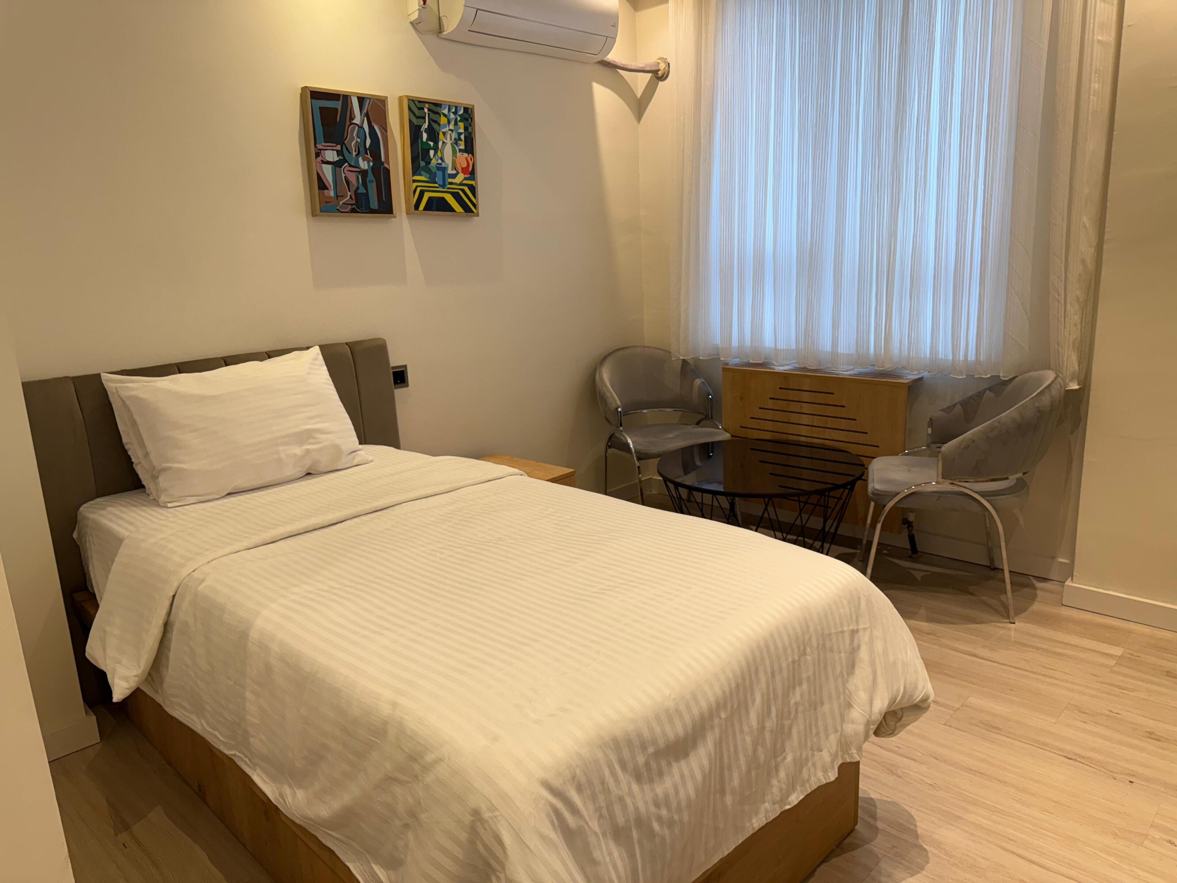 Una cama bien hecha con ropa blanca, una mesita redonda, dos sillas y dos cuadros abstractos en la pared.