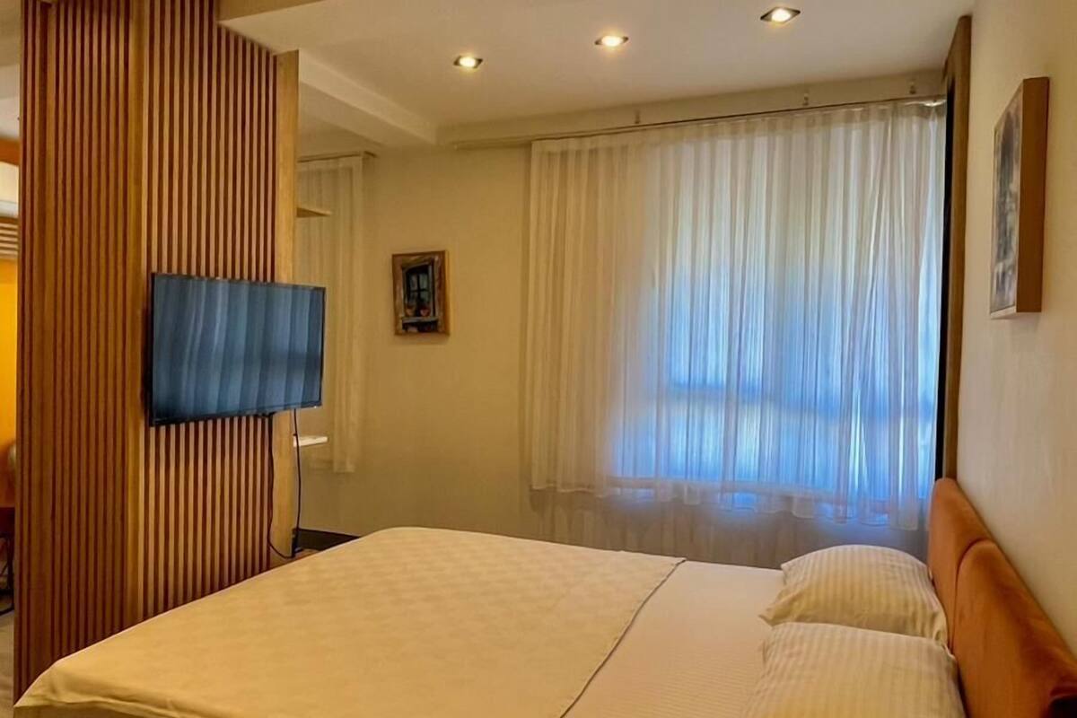 A hotel room with a large bed, a television, a wooden panel wall, and a window with curtains.