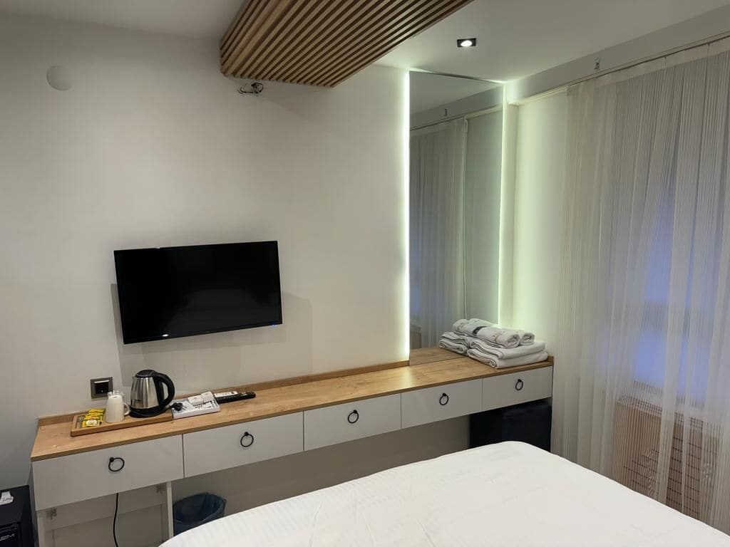 A modern hotel room with a flat-screen TV mounted on the wall, a wooden bedside table with a kettle, a remote, and a folded towel.