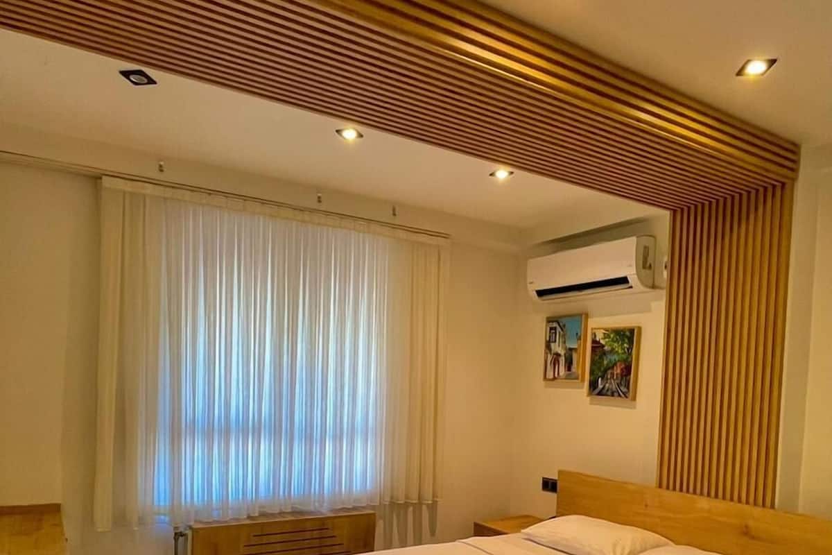 A modern hotel room with a wooden bed frame, a wooden desk, and a large window with white curtains.