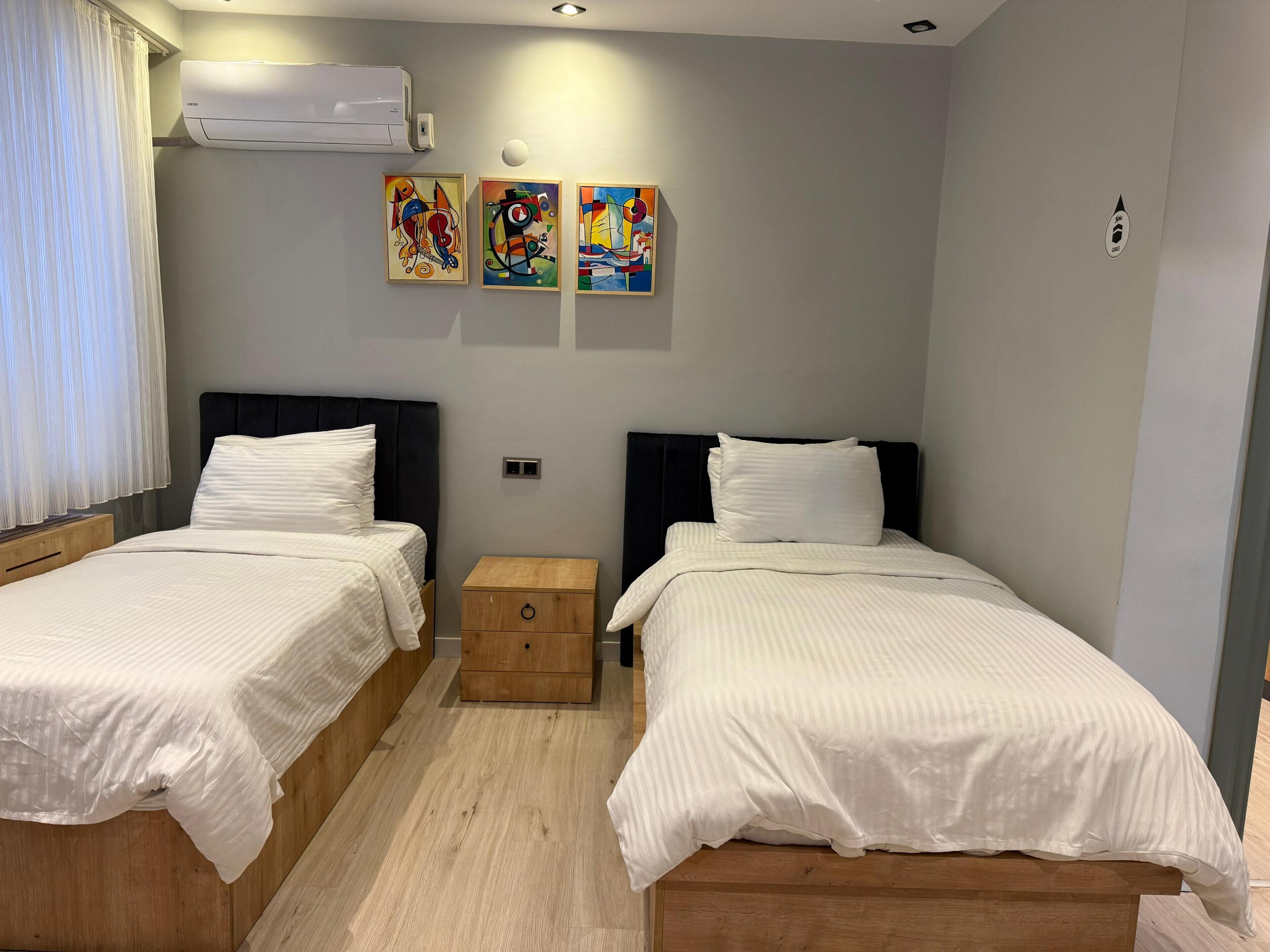 Une chambre double avec deux lits, chacun doté de draps blancs et d’une tête de lit noire. Trois tableaux abstraits aux couleurs vives sont accrochés au mur au-dessus des lits.