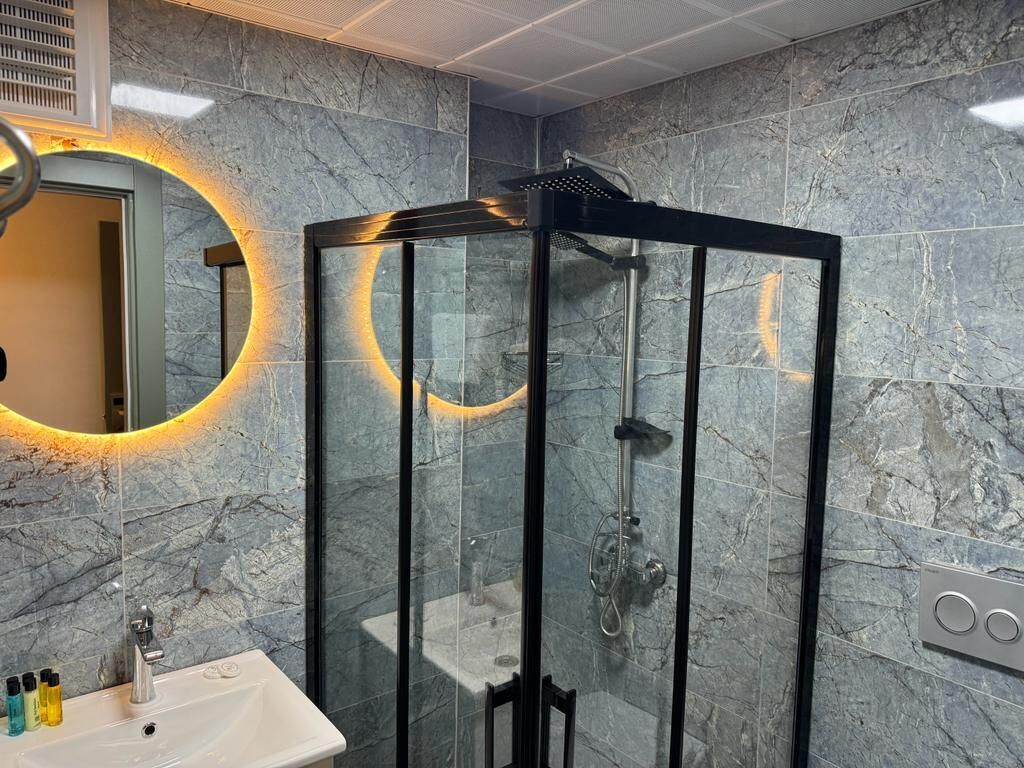 A modern bathroom with a glass-enclosed shower, marble walls, and a round mirror with a warm light.