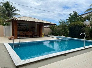 A rectangular swimming pool with a tiled deck, a small covered seating area with chairs, and a palm tree in the background. - Casa na Ilha - 100 metros da Praia  (Vera Cruz)