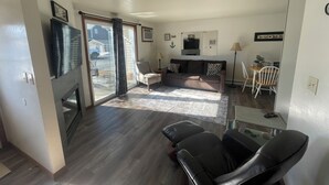 Een moderne woonkamer met een bank, een salontafel en een televisie. Er is een schuifdeur die leidt naar een terras met tuinmeubilair.