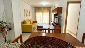 Ein Wohnzimmer mit gelbem Sofa, einem Esstisch mit Stühlen, einem Fernseher und einer Küchenzeile mit Schränken.