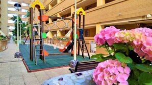 Ein Spielbereich für Kinder mit Klettergerüsten und Rutschen, umgeben von einem mehrstöckigen Gebäude mit Balkonen.