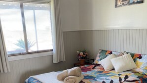 Un dormitorio con cama, ventana con persianas, mesita de noche con lámpara y un cuadro en la pared.