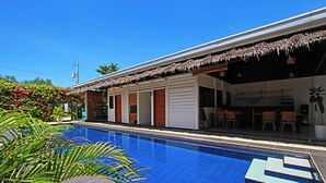 Una piscina con una estructura de techo de paja, rodeada de exuberante vegetación y un cielo azul despejado.