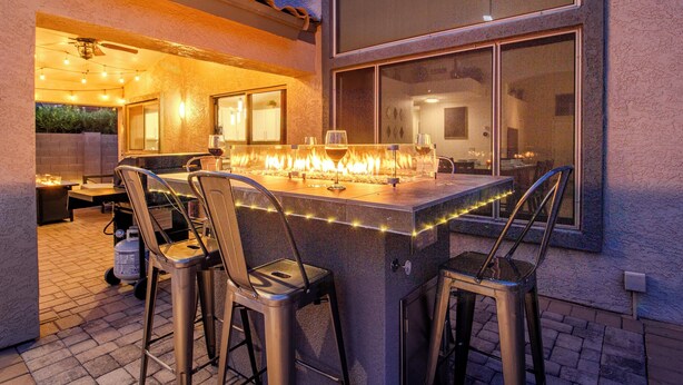 Un área de bar al aire libre con una chimenea, taburetes de bar y vistas al interior a través de un ventanal.