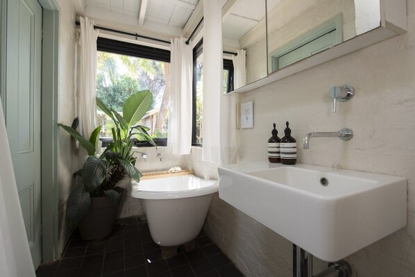 Un baño pequeño con un lavamanos blanco, un inodoro y una bañera. Hay plantas en macetas y una ventana que deja entrar luz natural.