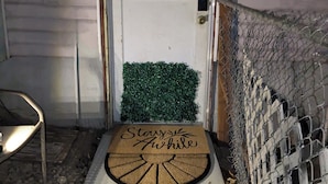 Una puerta de entrada con felpudo que dice “Stay & Awake”, un adorno decorativo sobre la puerta, un tapete de césped artificial verde y una alfombra estampada que conduce a la puerta.