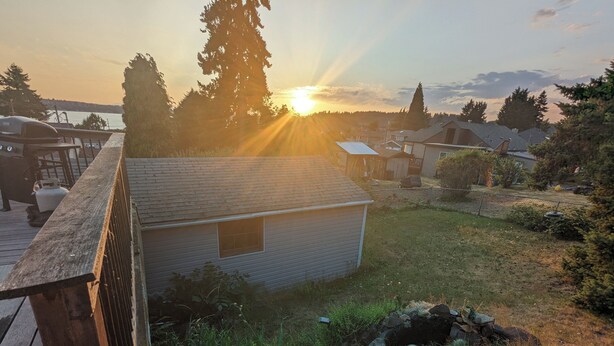 Una pequeña casa con terraza de madera, rodeada de vegetación y vista al atardecer sobre el agua.