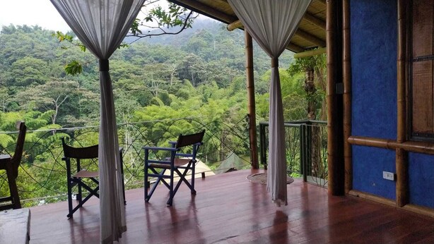 Terraza de madera con dos sillas y una mesa, con vistas a un bosque verde y frondoso.