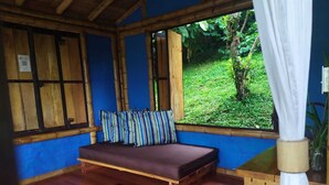 Una habitación con piso de madera, un sofá marrón con almohadones a rayas, una ventana con vista a la vegetación y un techo de bambú.
