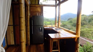Cocina compacta con refrigerador, un lavamanos y una mesa de madera, con vista a un paisaje verde y exuberante.