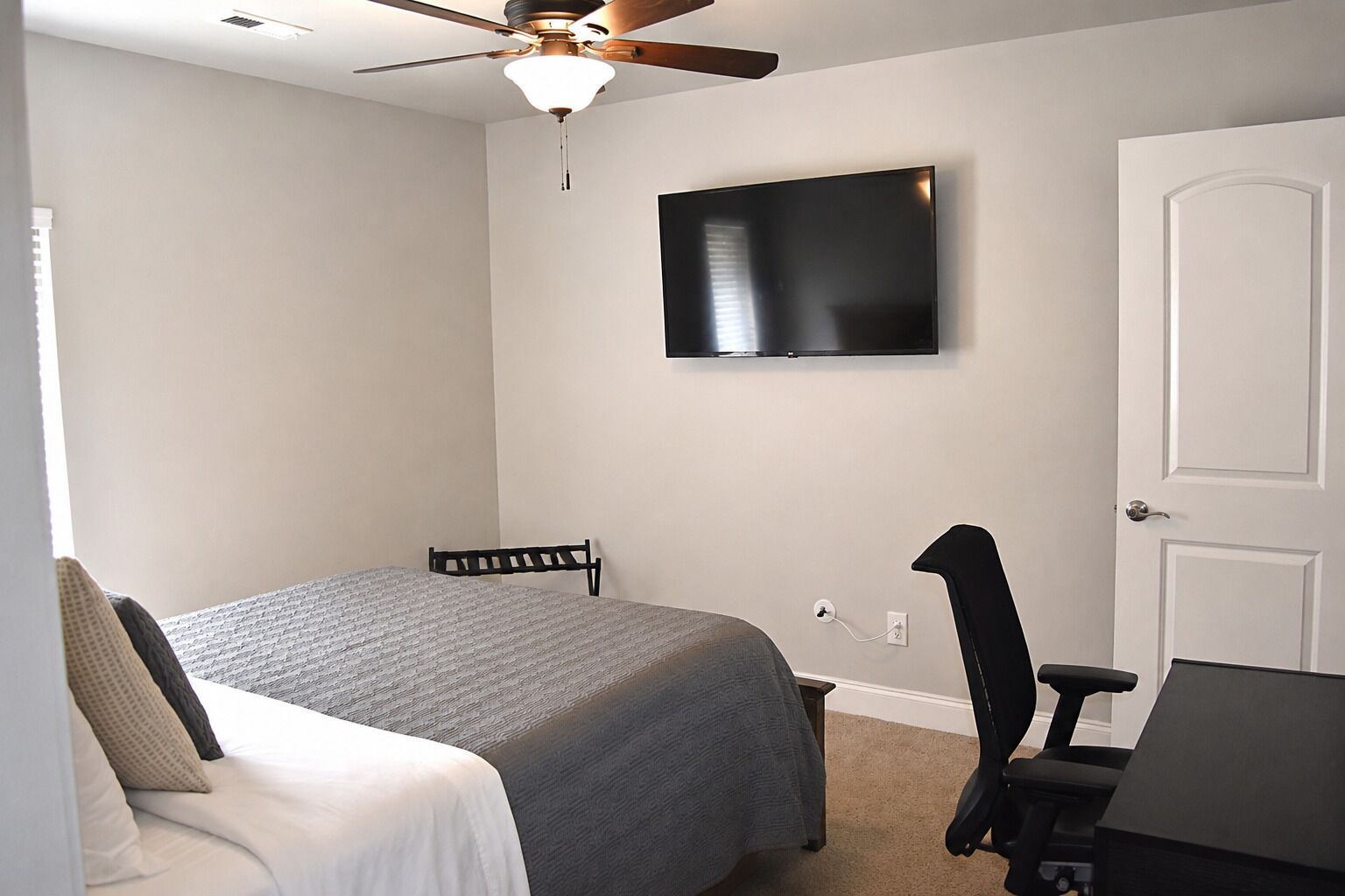 Ein Schlafzimmer mit einem Bett, einem Schreibtisch, einem Stuhl, einem Deckenventilator und einem an der Wand montierten Fernseher.