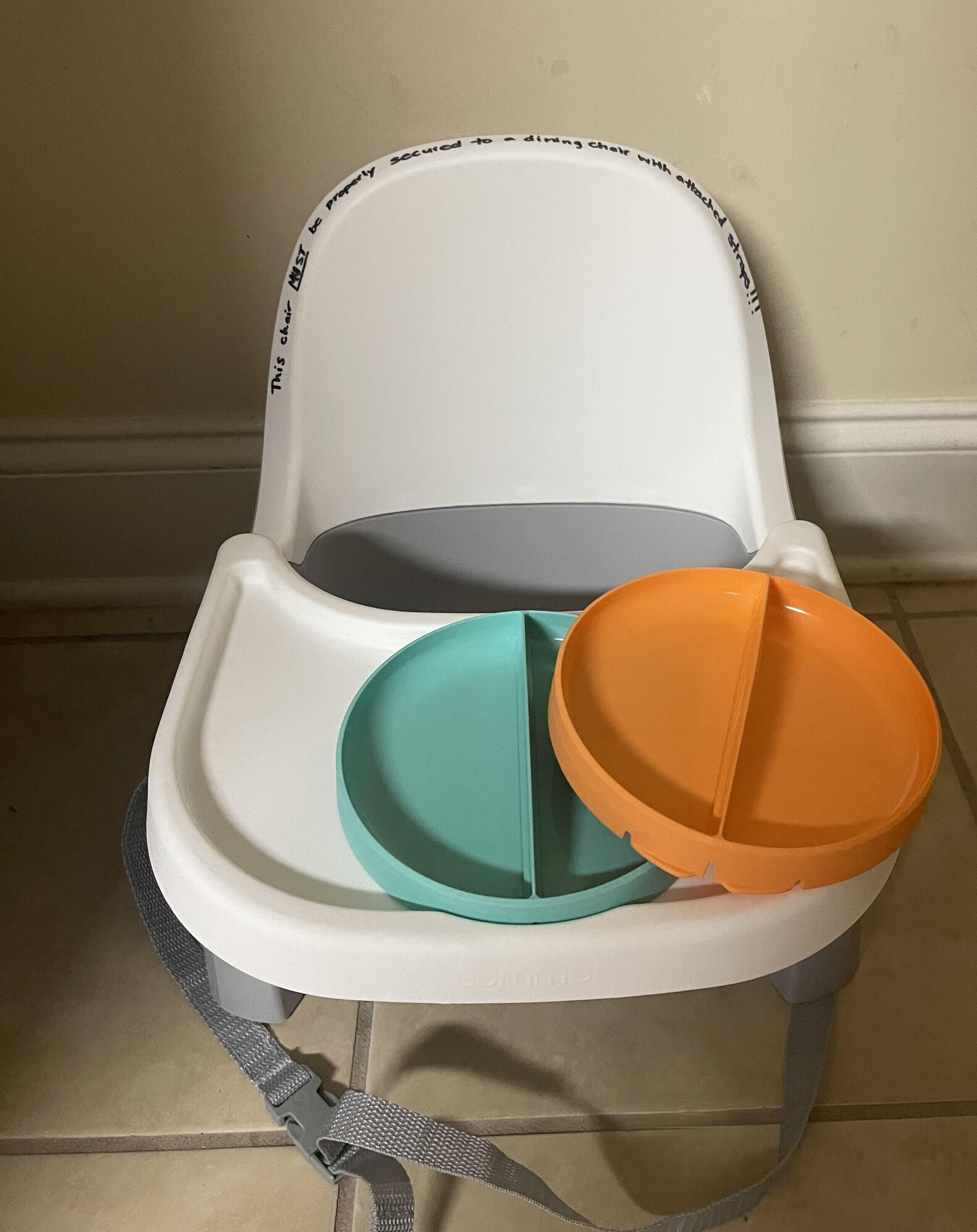 A high chair with a tray and two divided plates, one teal and one orange, against a plain wall.