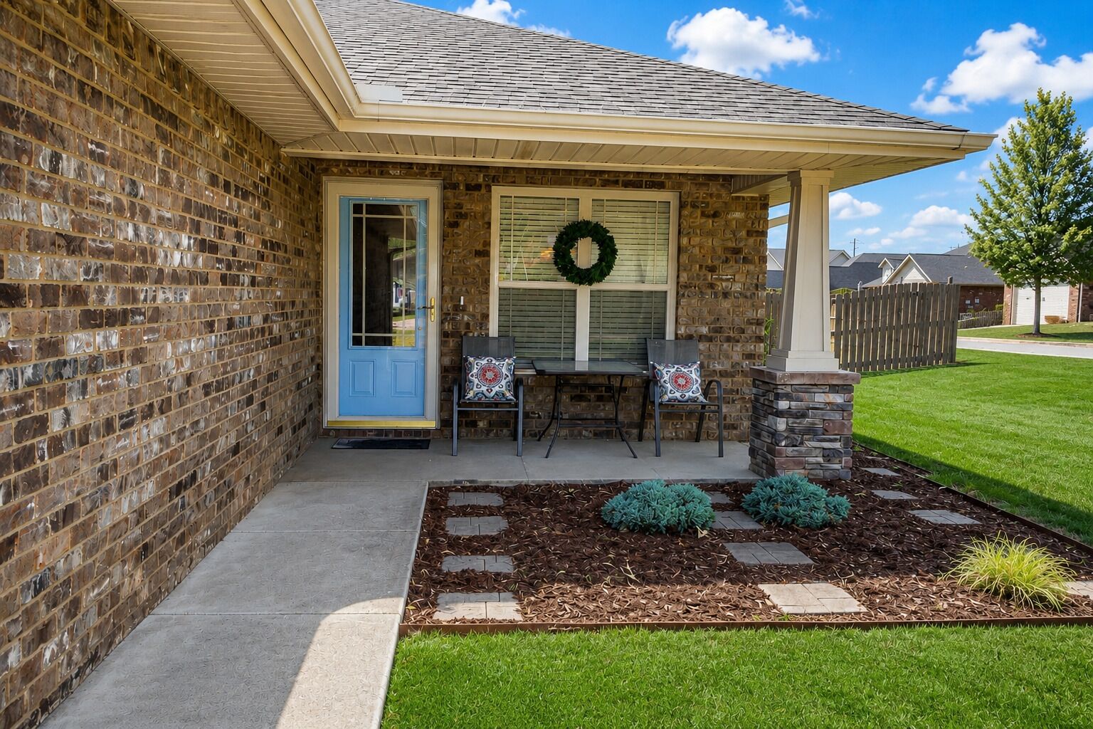 A brick house with a blue door, a covered porch, a stone pillar, and a wreath on the window.