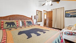 A room with a large bed featuring a bear-themed quilt, a ceiling fan, a wooden door, and a television on the wall.