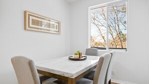 A dining area with a white marble table, four grey upholstered chairs, a framed artwork on the wall, and a window with a view of bare trees. - The Bear House - Luxury Big Bear Home | Game Room, Modern (Big Bear City)