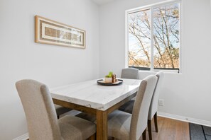 Een eetgedeelte met een witte marmeren tafel, vier grijs gestoffeerde stoelen, een lijstwerk met kunst en een raam met uitzicht op kale bomen.