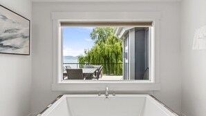A bathroom with a bathtub, a window with a view of a patio and greenery, and a framed picture on the wall.