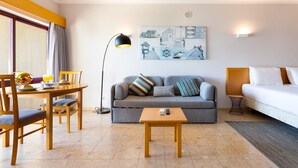 Ein modernes Hotelzimmer mit Essbereich, einem grauen Sofa, einem kleinen Tisch und einem Bett.